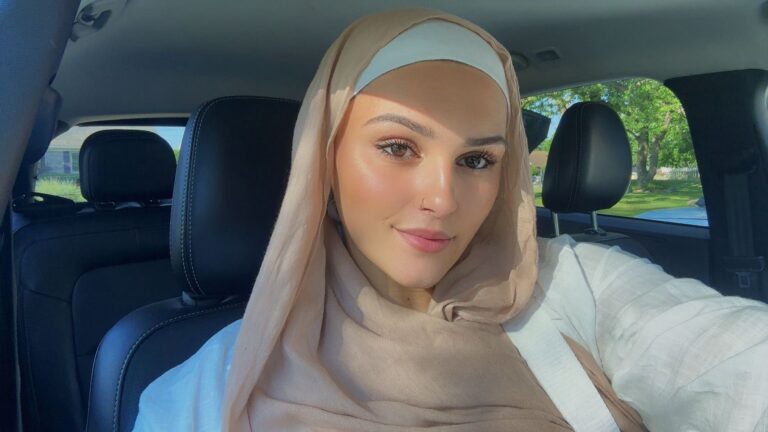 A woman wearing a beige hijab and white top sitting inside a car.