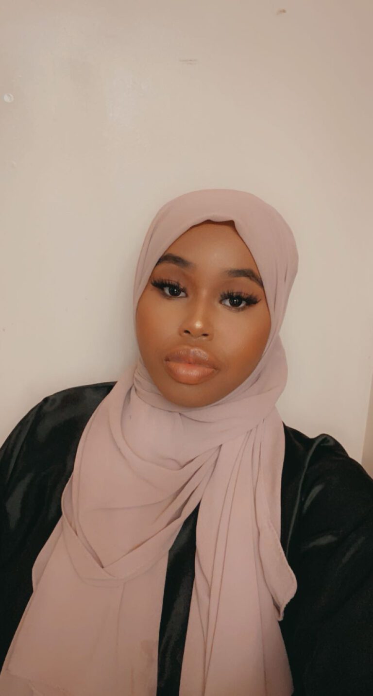 A woman wearing a light pink hijab and makeup, looking directly at the camera.