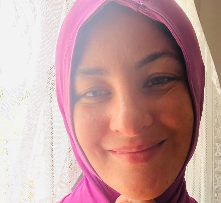 Smiling person in purple headscarf near window.
