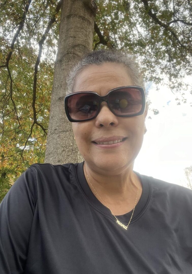 Smiling woman wearing sunglasses outdoors with trees in the background.