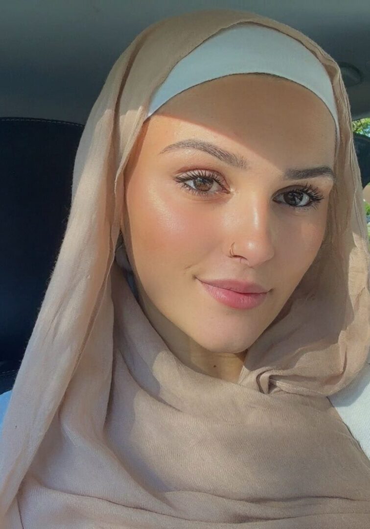 A woman wearing a beige hijab and white top sitting inside a car.