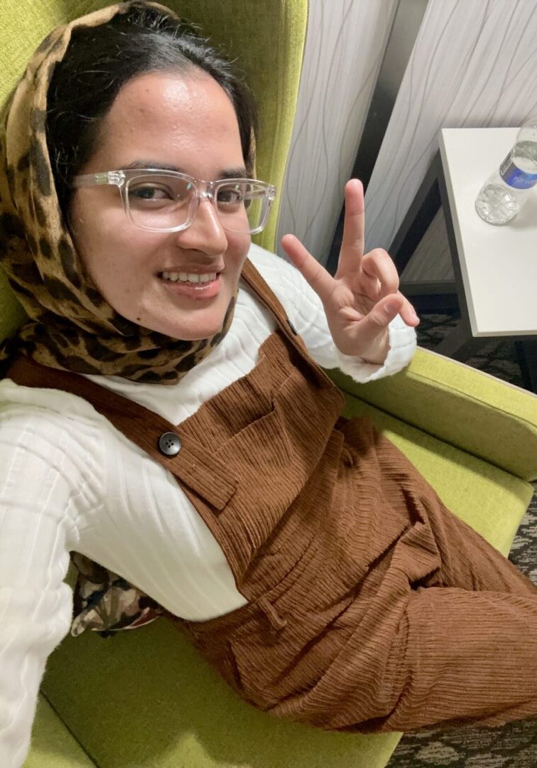 Woman sitting, smiling, giving peace sign.