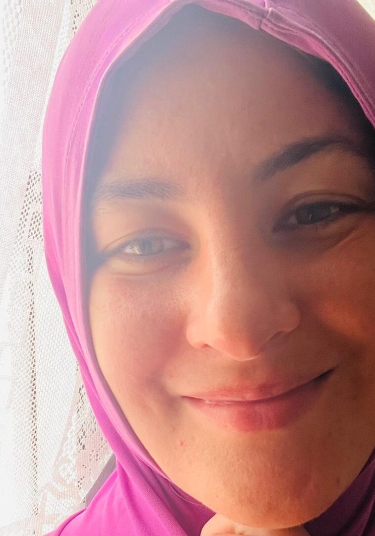 Smiling person in purple headscarf near window.